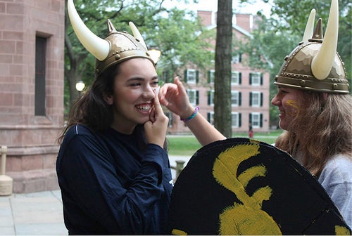 Two female students in viking helmet, while one helps the other apply facepaint.