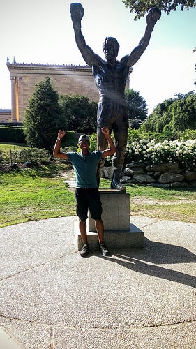 Author Abdul standing with the Rocky Balboa statue, raising his arms trimphantly in imitation.
