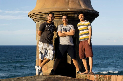 Three men lean against a large pillar by the ocean.