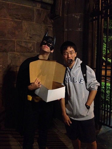&quot;Batman&quot; with a box of donuts, posing with a student with a whole donut hanging out of his mouth.