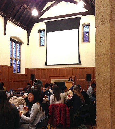 A student speaking from a podium at the head of the dining hall.