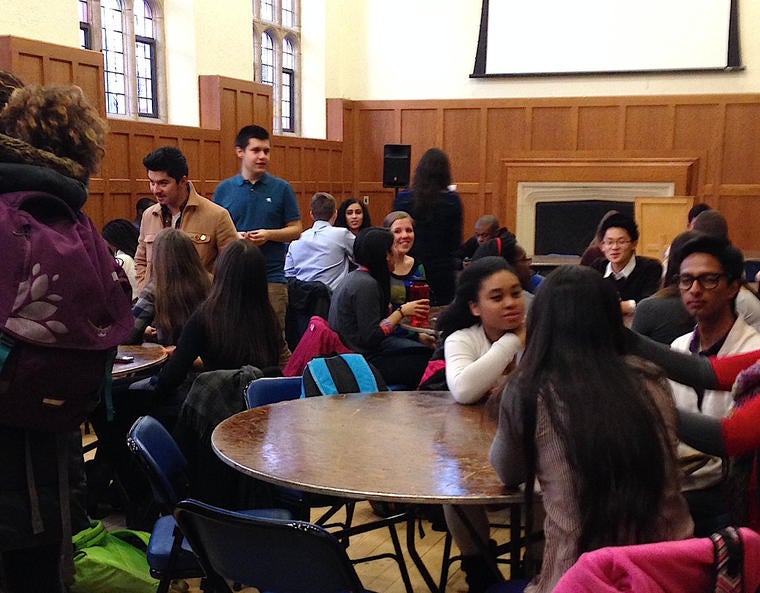 A diverse group of students in conversation with each-other, seated together at small tables.