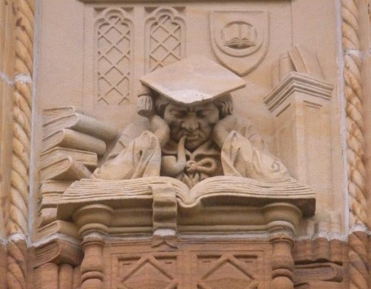 A carving of a scholar in robes and mortarboard, sleeping with his head propped up above an enormous book.