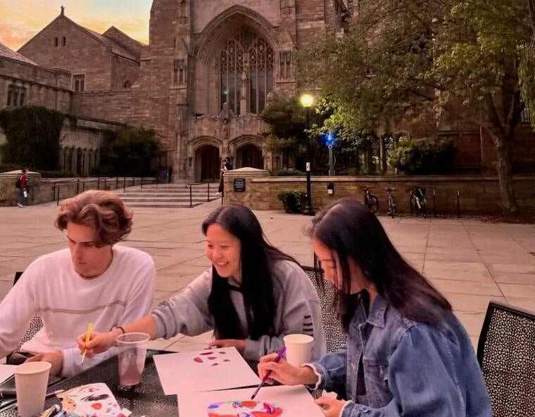 Me sitting a table with two friends and painting outside of Sterling Memorial Library.