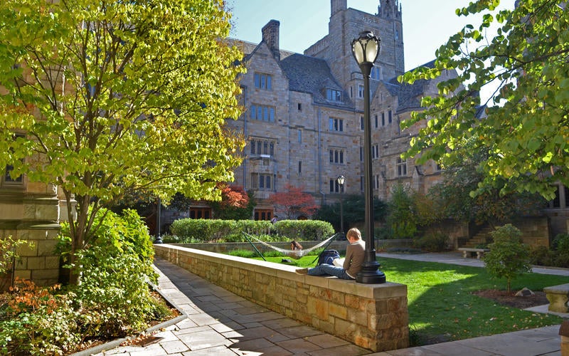 A student sits on a wall to read while another lounges in a hammock on a Residential College lawn at Yale